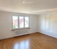 Empty room with wooden flooring, white walls, and large windows offering a view of neighboring houses.