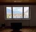 Empty room with wooden flooring, large windows offering a scenic mountain view, a table lamp, and a television on a stand.