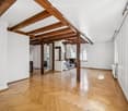 Bright, open corridor with exposed wooden beams and herringbone parquet flooring. White walls, large windows, and visible kitchen area in the background.