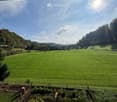 View from a balcony or terrace overlooking a lush green field with scattered trees and distant houses under a clear blue sky.