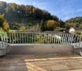 Balcony with wooden decking, white metal railing, overlooking a green landscape with trees and houses.