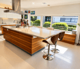 Modern kitchen with a large island, white countertops, wooden cabinetry, and four bar stools. Large windows provide natural light, and the space features a sleek, minimalist design.
