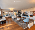 Spacious living room with hardwood floors, large windows, and a fireplace. Features a white sectional sofa, coffee table, ottoman, armchairs, and a dining area with a round table and chairs. Decor includes artwork, bookshelves, and decorative candles.