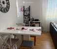 A modern dining area with a white table, floral placemats, and four chairs, including a wooden chair with a cushion. A sofa is visible to the right, and a small shelf holds kitchen items like a teapot, cups, and a coffee maker. A large wall clock and a fr