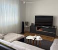 Modern living room with a grey sofa, coffee table, TV stand, and a small round table with a lamp and fan. The room features wooden flooring and large windows with white curtains.