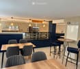 Modern pizzeria interior with wooden tables, upholstered chairs, bar stools, and a visible kitchen area with an oven and a sink.