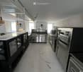 Commercial kitchen with stainless steel appliances, including a refrigerator, oven, and sink, along with a counter and pantry. The space features pendant lighting and large windows.