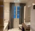 Modern bathroom with white tiles, a bathtub, toilet, sink, and a wooden vanity. Features include a large window with blinds, a decorative vase with flowers, and a green bath mat.