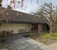 Outdoor patio area with stone tiles, wooden furniture including a table and chairs, small garden plants, and a view of a house with a tiled roof.