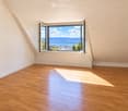 Empty room with wooden flooring and white walls, featuring a large window offering a scenic view of a lake and mountains.