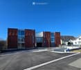 Modern apartment building with red facade, glass windows, parking area in front