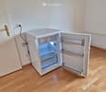 A small, white refrigerator with its door open, showing empty shelves and compartments. The room has wooden flooring and white walls.