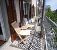 Balcony with a grey sofa, wooden folding chair, small table, patterned rug, and potted plants, overlooking a cityscape.
