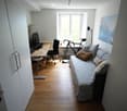 A compact, well-lit room with a sofa, desk, and bicycle. The space features a wooden floor, large windows, and a mix of modern and cozy furnishings.