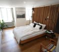 Cozy bedroom with wooden flooring, a bed with multiple pillows and a duvet, a nightstand with books, a colorful chair, a potted plant near the window, and a picture frame on the wall.