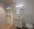 White tiled walls, glass walk-in shower, vanity with sink and drawers, and a toilet. The shower area has tiled flooring.