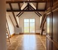 An empty attic space with exposed wooden beams, a large window with decorative ironwork, and a patterned wooden floor.