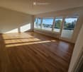 Empty room with wooden flooring and large windows offering a view of the city and trees. The room is unfurnished with natural light streaming in.