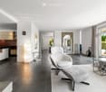 Modern living room with a grey sofa, white armchair, ottoman, coffee table, and a sleek kitchen area with a bar counter and bookshelf. Large windows and a mirror add brightness and depth to the space.