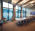 Modern conference room with large windows, a long conference table surrounded by office chairs, side chairs, a small round table, and a screen mounted on the wall.