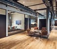 Modern office space with wooden flooring, glass partitions, leather chairs, small tables, and a screen in the background.