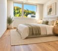 Modern bedroom with a large bed, nightstands, lamps, a rug, and a potted plant near the window. The room features wooden flooring and large windows with a scenic view.