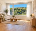 Bright living room with large windows offering a scenic view, featuring a white sofa, coffee table, side table, floor lamp, sideboard, and decorative plants.