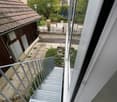 Balcony with metal stairs leading down to a small outdoor area with concrete flooring, greenery, and a wooden shed.