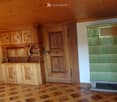 A cozy room with wooden walls, a patterned wooden floor, and a green-tiled fireplace. Wooden cabinets and shelves are mounted on the wall, and a door is visible.