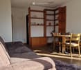 A modern living room with a grey sofa, wooden dining table, four chairs, and a wooden shelf with decorative items.