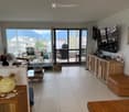 Bright living room with large sliding glass doors leading to a balcony, offering a scenic mountain view. The room features a sofa, coffee table, ottoman, chairs, a television, cabinets, and decorative items including a plant and a ceramic pig figurine.