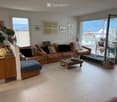 Bright living room with large windows and a view, featuring a brown leather sofa, glass coffee table, ottoman, and decorative plant on a side table.