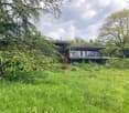 A two-story wooden house with a raised ground floor, surrounded by lush greenery and trees. The house features a balcony with outdoor seating and a chimney on the roof.