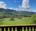 Balcony overlooking a lush green valley with rolling hills, scattered trees, and distant mountains under a partly cloudy sky.