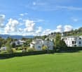 A well-maintained lawn with a hedge border, surrounded by residential buildings and trees, with a scenic mountain backdrop under a partly cloudy sky.