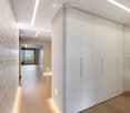 Modern hallway with light wooden flooring, white built-in wardrobes, recessed lighting, and a textured brick accent wall. The hallway leads to a room with large windows and a balcony view.