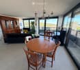Bright living and dining area with large windows offering a scenic view, featuring a sofa, dining table with chairs, television, and a wooden cabinet.