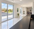 Modern, minimalist hallway with large glass windows, polished marble floors, recessed lighting, and a sleek black table with a stool. Potted plants and artworks adorn the walls.