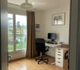 Bright, modern home office with wooden flooring, large windows offering a view, a desk with a computer, a black office chair, a white storage unit, and a hanging lamp.