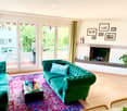 Modern living room with teal sofa, glass coffee table, fireplace, large windows, and outdoor seating area visible through glass doors.