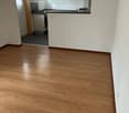 Empty room with wooden flooring and white walls, featuring a doorway to a small kitchen area with cabinets and a countertop.