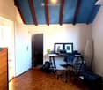 A modern, furnished room with wooden flooring, a desk with a laptop, a chair, and framed artwork on the wall. The room features exposed wooden beams and a slanted ceiling.
