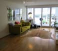 Bright living room with large windows and a balcony, featuring a green sofa, coffee table, floor lamp, and decorative mirror.