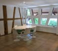 Modern dining room with a white round table, six white chairs, pendant lights, and large windows with wooden shutters.