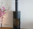 Modern black wood-burning stove with a tall chimney, placed on a glass platform in a room with wooden flooring and white walls. A vase with pink flowers is visible to the left.
