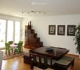 Bright living and dining area with large windows and sliding doors leading to a balcony. Features a grey sofa, coffee table, dining table with chairs, wooden cabinet, decorative vases, picture frames, and a large potted plant.