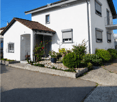 Two-story white house with a tiled roof, balcony, and small garden area with potted plants.