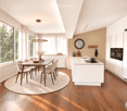 Modern kitchen with white cabinets, wooden flooring, and a central island. A dining area with a wooden table and white chairs is adjacent, featuring a round rug and a vase with flowers. Large windows provide natural light, and a pendant light hangs above