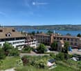 A multi-story residential building with a tiled roof, surrounded by greenery and parked cars, overlooking a lake and distant cityscape.