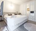 Modern bedroom with a large white bed, pillows, a wardrobe, a chair, a screen, a table, and a rug.
